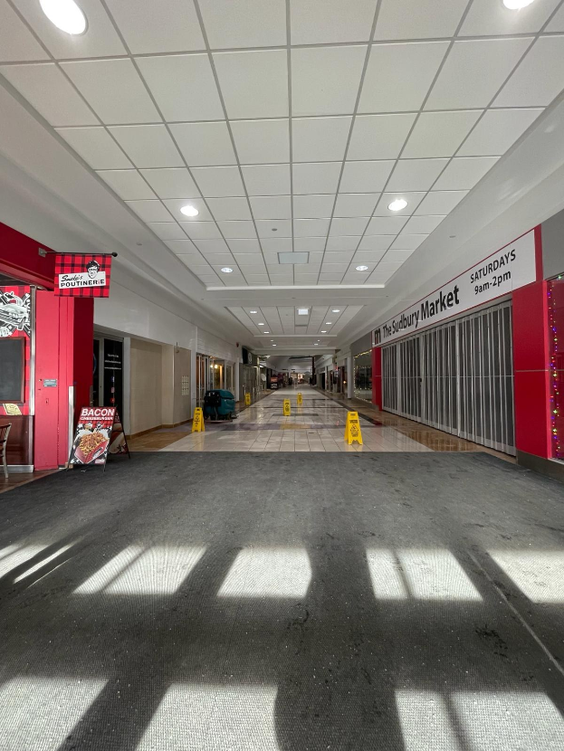 Innenansicht eines Einkaufszentrums mit Deckenbeleuchtung, einem Stuhl und einer Tafel, auf der "Sydbury Market - Vorher und Nachher" steht, links, und verstreuten Gegenständen rechts und auf dem Boden.