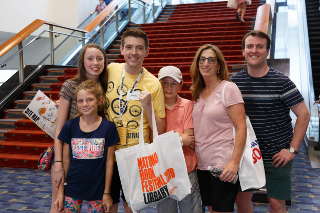 Eine Gruppe strahlender Menschen mit Taschen steht zusammen vor einer Rolltreppe auf dem Nationalen Buchfest 2019, wobei Treppen, Geländer und eine Wand im Hintergrund zu sehen sind.