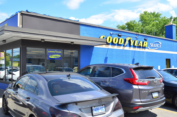 Außenansicht einer Goodyear Select Autohandlung mit geparkten Fahrzeugen, Gebäudeschild, Bäumen und einem bewölkten Himmel.