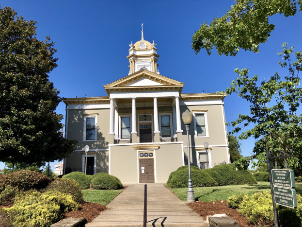 Großes Backsteingebäude mit einem Glockenturm, mehreren Fenstern, Türen und Säulen, umgeben von Straßenmasten, Laternen, einem Schild, Bäumen, Gras und einem bewölkten Himmel, als das wiedereröffnete alte Rathaus bezeichnet.