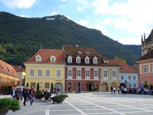 Gruppe von Menschen, die durch einen Stadtplatz mit Gebäuden, Fenstern, Laternen und Topfpflanzen geht, mit einem Hügel voller Bäume und einem bewölkten Himmel im Hintergrund.