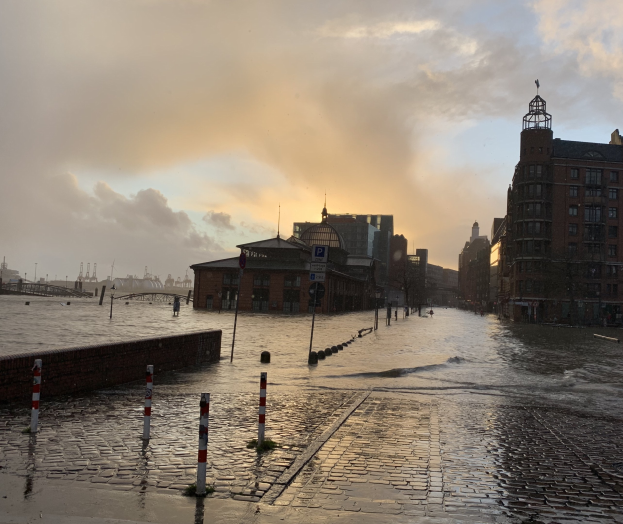 Überschwemmte Straße in Hamburg, Deutschland, mit Wasser, das die Straße, Pfosten, Schilder, Gebäude mit Fenstern, eine Brücke und einen bewölkten Himmel bedeckt.