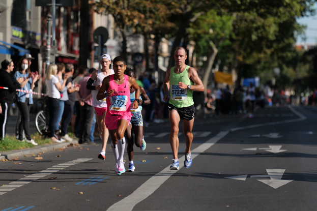 Gruppe von Menschen beim Laufen in einem Stadtmarathon mit Zuschauern auf der linken Seite, unscharfer städtischer Hintergrund mit Bäumen, Gebäuden und einem Fahrrad.