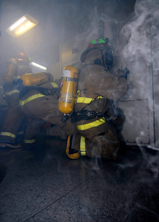 Feuerwehrleute in Schutzausrüstung mit rauchgefülltem Raum und beleuchteter Wand im Hintergrund.