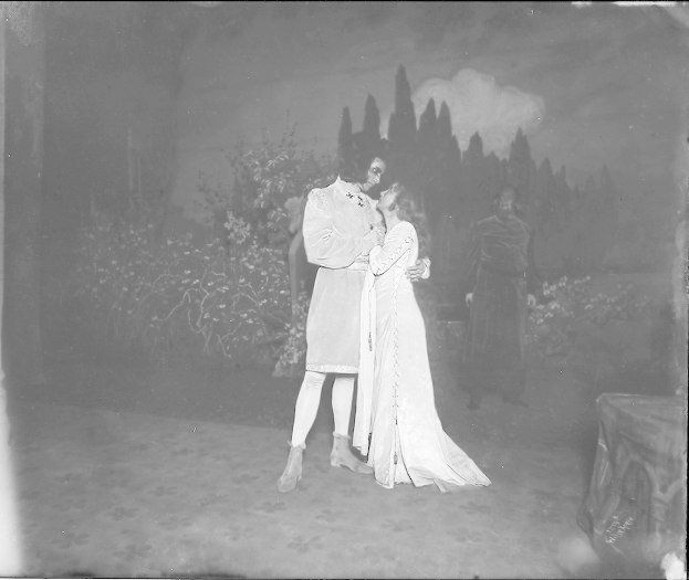 Ein Schwarz-Weiß-Foto eines Mannes und einer Frau, die sich auf einer Bühne umarmen und küssen, mit ein paar Menschen, Pflanzen und Bäumen im Hintergrund.