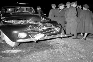 Eine Schwarz-Weiss-Szene eines beschädigten Autos am Strassenrand, mit einer Gruppe von Menschen drumherum, die besorgt aussehen.