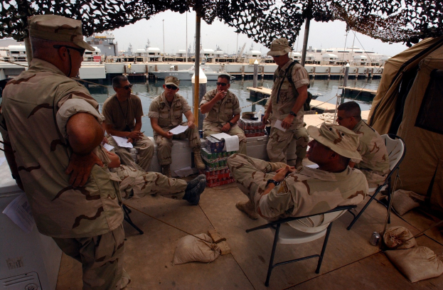 Gruppe von Männern mit Mützen und Schwimmbrillen, die um einen Tisch auf einem Boot sitzen, mit Taschen auf dem Boden und einem Zelt auf der rechten Seite, vor dem Hintergrund von Booten, Pfählen, Gebäuden und einem klaren blauen Himmel.