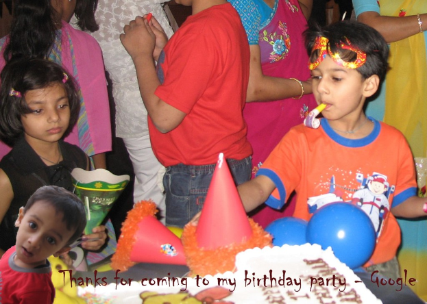 Kinder um einen Tisch mit einem Kuchen, Luftballons und Partyhüten versammelt, mit weiteren Personen im Hintergrund.