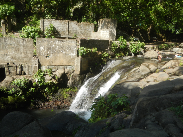 Ein kleiner Wasserfall ergießt sich über eine felsige Klippe in einem üppigen grünen Wald, mit Menschen, die im Wasser schwimmen und einer Wand im Hintergrund.