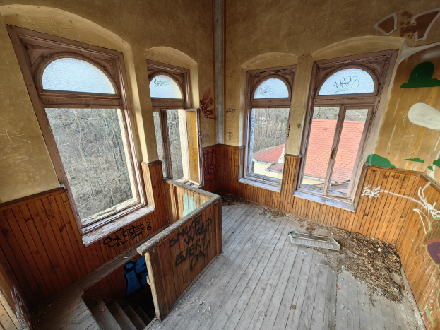 Ein verlassenes Gebäude mit Graffiti an den Wänden und Fenstern, einem Holzfußboden, einer Treppe und verstreuten Gegenständen, mit Bäumen und einem Haus im Hintergrund.