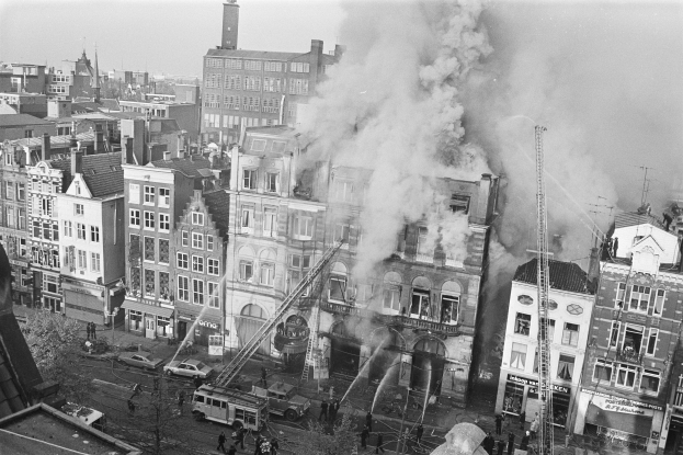 Ein Schwarz-Weiß-Foto einer Stadtstraße mit einem Feuer in der Mitte, umgeben von Gebäuden mit Fenstern, Fahrzeugen und einigen Menschen, mit Rauch, der aus den Gebäuden aufsteigt und der Himmel im Hintergrund sichtbar ist.