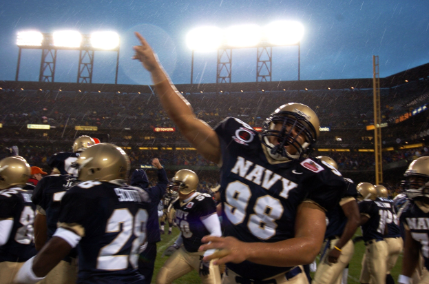 Navy-Football-Spieler feiern ein Touchdown auf einem regennassen Feld, ihre Uniformen und Helme sind sichtbar, mit Zuschauern und Stadionbeleuchtung im Hintergrund.