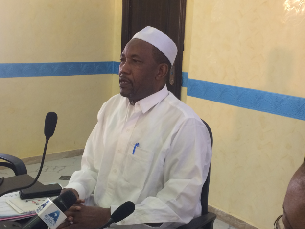 Ein Mann in einem weißen Gewand und Hut sitzt an einem Tisch mit Mikrofonen, spricht, während Papiere und Gegenstände auf dem Tisch zu sehen sind, mit einer anderen Person mit Brille rechts von ihm, in einem Raum mit einer Wand und einer Tür dahinter.