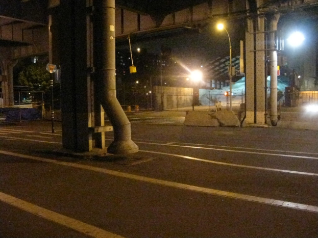 Eine Straße mit Säulen, Rohren, Pfählen, Laternen, einem Schuppen, einem Zaun, Bäumen und Gebäuden im Hintergrund.