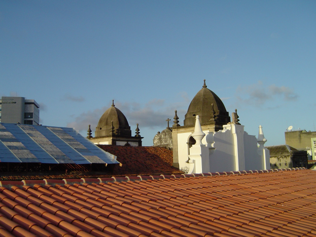 Stadtansicht mit mehreren Gebäuden im Vordergrund unter einem blauen Himmel, mit Solarpanelen auf einem Dach, die den Einsatz erneuerbarer Energien anzeigen.
