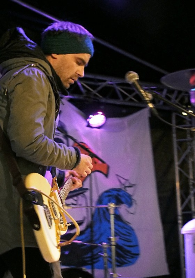 Ein Mann in einer Jacke und einer Mütze spielt eine E-Gitarre auf der Bühne mit einem Mikrofon davor, einem Schlagzeug zu seiner Rechten und Scheinwerfern und einem Banner im Hintergrund.