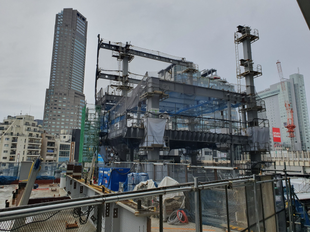 Großer Baustelle in einer Stadt mit einem teilweise errichteten Kraftwerk, umgeben von einem Metallzaun, Fahrzeugen, Strommästen, einer Plane und einem bewölktem Himmel.
