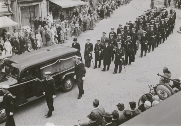 Schwarz-weißes Bild einer Beerdigungsprozession mit einem Leichenwagen, der den Körper eines Mannes trägt, umgeben von Menschen, einige halten Waffen, mit Fahrzeugen und Gebäuden im Hintergrund.