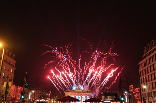 Eine belebte Stadtstraße in Berlin an Silvester, voller Menschen, Fahrzeuge und Gebäude, erleuchtet von Feuerwerk und Gebäudelichtern, die eine festliche Atmosphäre schaffen.