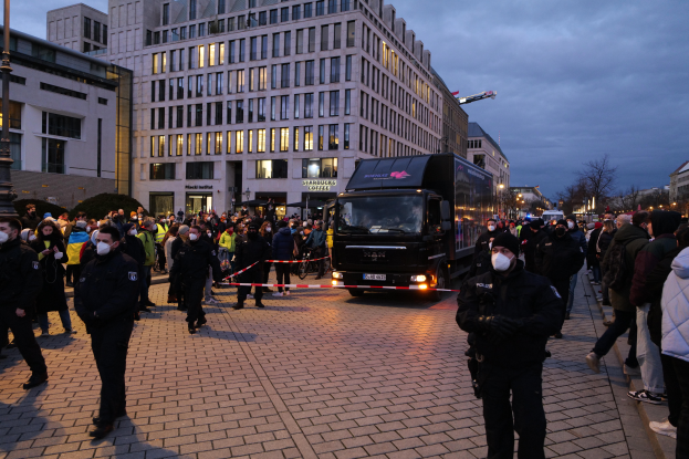Eine Gruppe von Menschen steht vor einem Lkw auf einer Straße, umgeben von Gebäuden, Laternen, Bäumen und einem bewölkten Himmel, wobei einige Mützen und Masken tragen und ein Band mit einem Pfahl im Vordergrund.