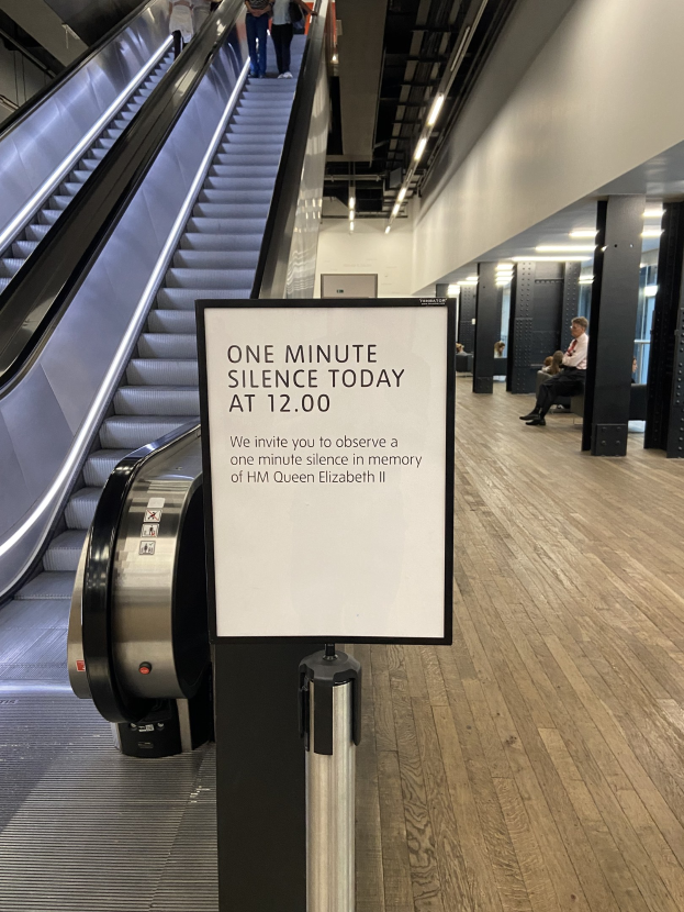 Eine Rolltreppe in einem Flughafen mit einem Schild, auf dem "Eine Minute Stille heute" steht, sowie ein paar Menschen darauf und an der Decke angebrachte Lampen im Hintergrund.
