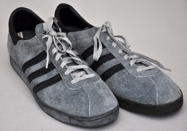 Ein Paar graue und schwarze Adidas Gazelle-Schuhe mit klassischem Design, schwarzen Akzenten und einer weißen Sohle, auf einer weißen Oberfläche platziert.