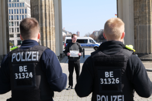 Drei Polizeibeamte stehen vor einem Gebäude, wobei ein Mann in der Mitte ein Papier hält, flankiert von Säulen, mit Fahrzeugen, Menschen, Bäumen, Gebäuden und Himmel im Hintergrund.