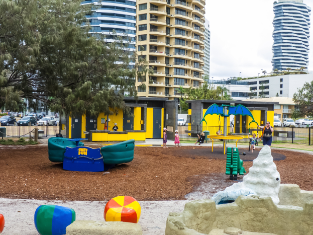 Ein Spielplatz in der Stadt mit Kindern auf Spielgeräten, umgeben von Bäumen und Zäunen, mit hohen Gebäuden und Fahrzeugen im Hintergrund, unter einem sichtbaren Himmel.