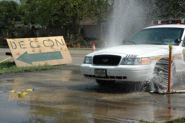 Ein Streifenwagen fährt durch eine Wasserlache, mit einem Schild, einer Verkehrskegel und anderen Gegenständen am Boden, mit Bäumen, Gebäuden und einem klaren blauen Himmel im Hintergrund.