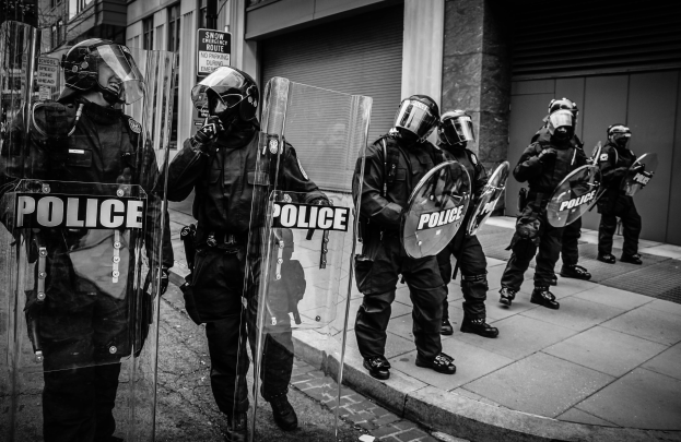Eine Gruppe von Polizisten in Helmen und mit Schilden mit Text darauf steht auf einem Bürgersteig vor Gebäuden mit Fenstern, Läden und Schildern in einem Schwarz-Weiß-Bild.