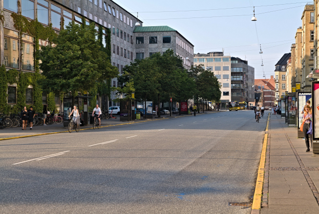Stadtstraße mit Fahrradfahrern, hohen Gebäuden, Bäumen, Laternen und Texttafeln, mit Himmel im Hintergrund.