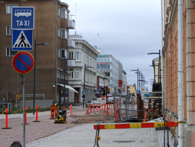 Eine Stadtstraße mit Gebäuden, Straßenlaternen, Verkehrsschildern, Verkehrszeichen, Baustellenabsperrungen, Kraftfahrzeugen, Absperrpoller, Bäumen und einer Baustelle mit Verkehrsschildern unter einem bewölkten Himmel.